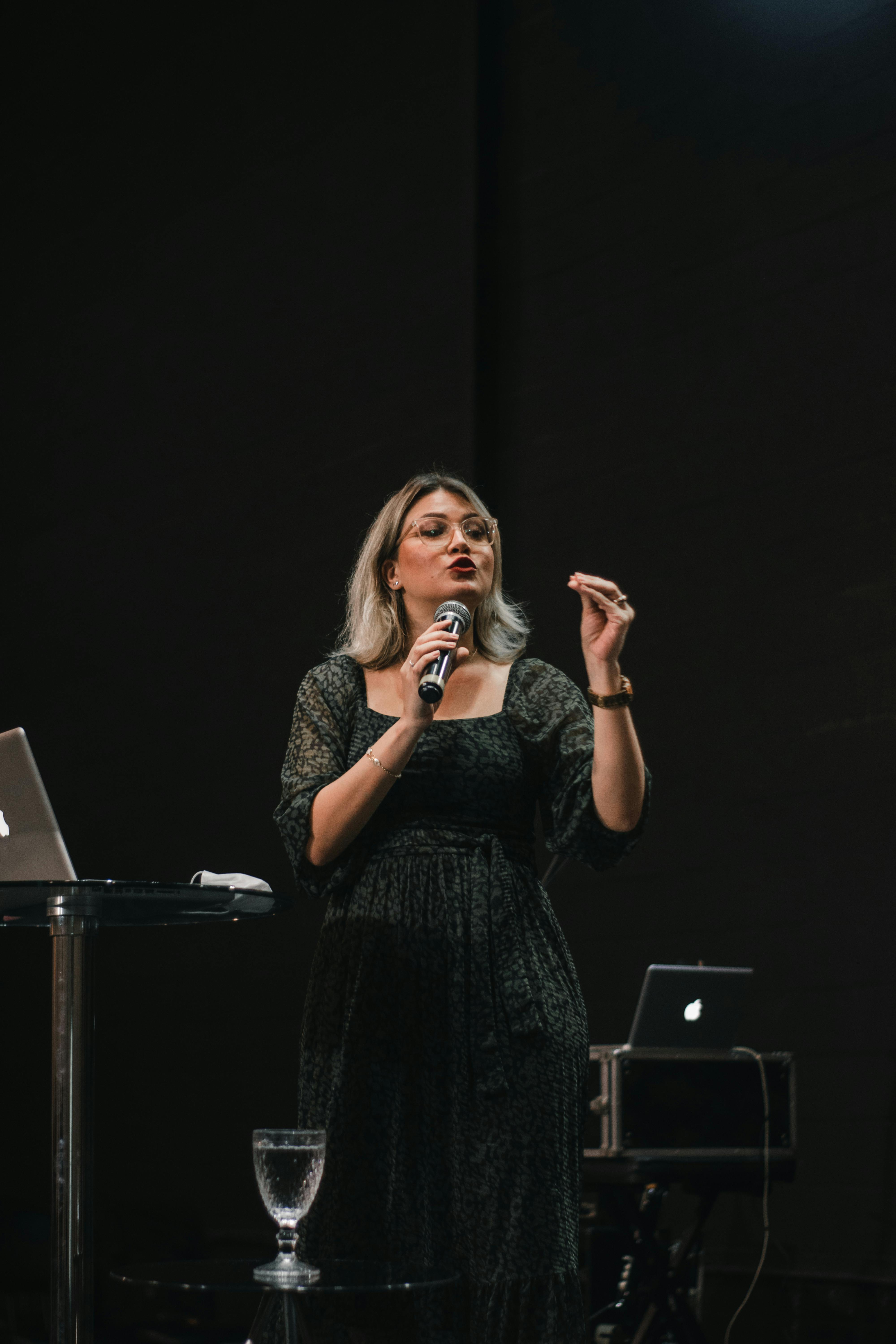 Woman with microphone giving a speech on a dark stage. Audience captivated by her performance.