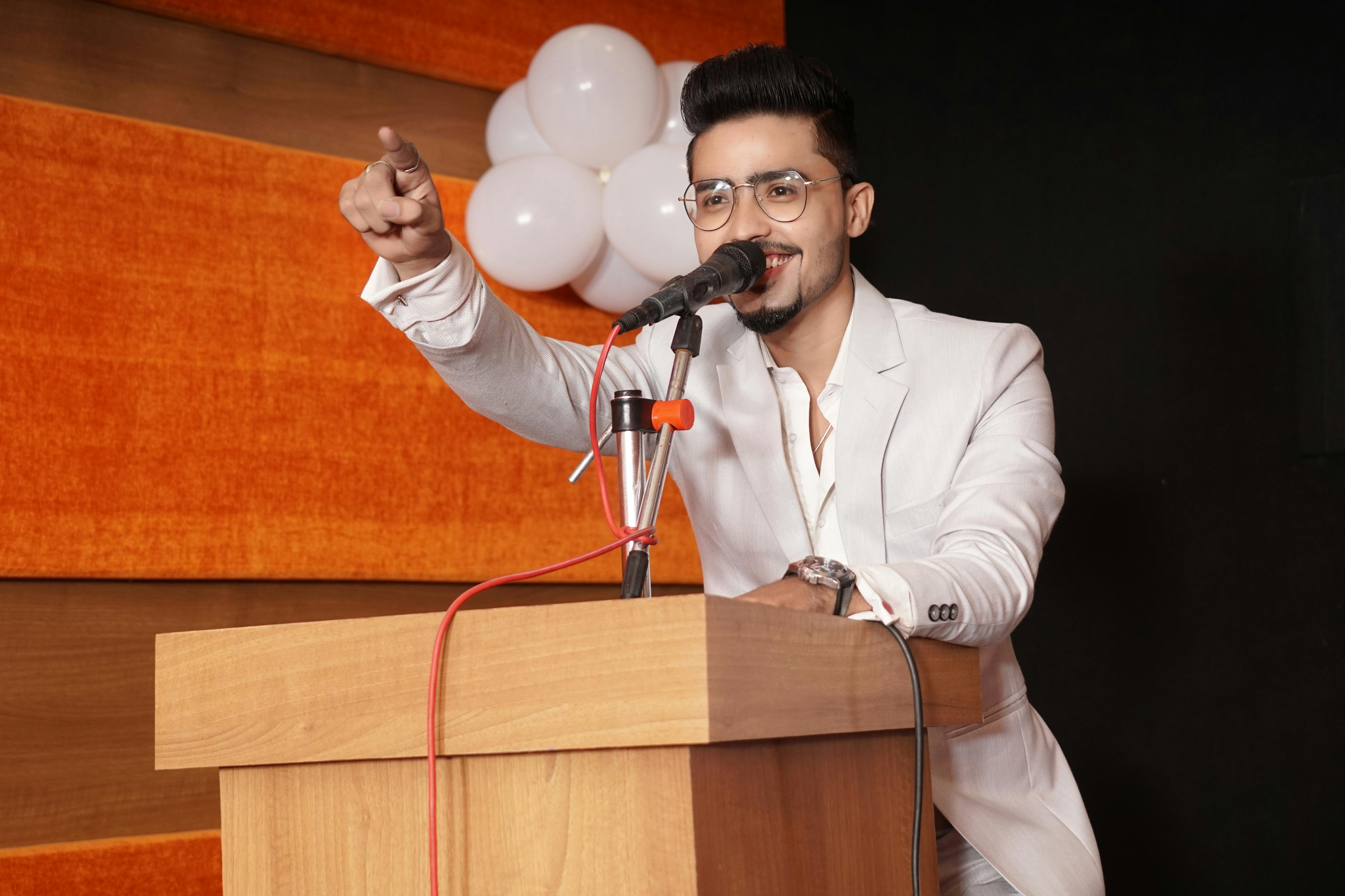 A smiling man in a white suit giving a speech at a podium with balloons in the background.